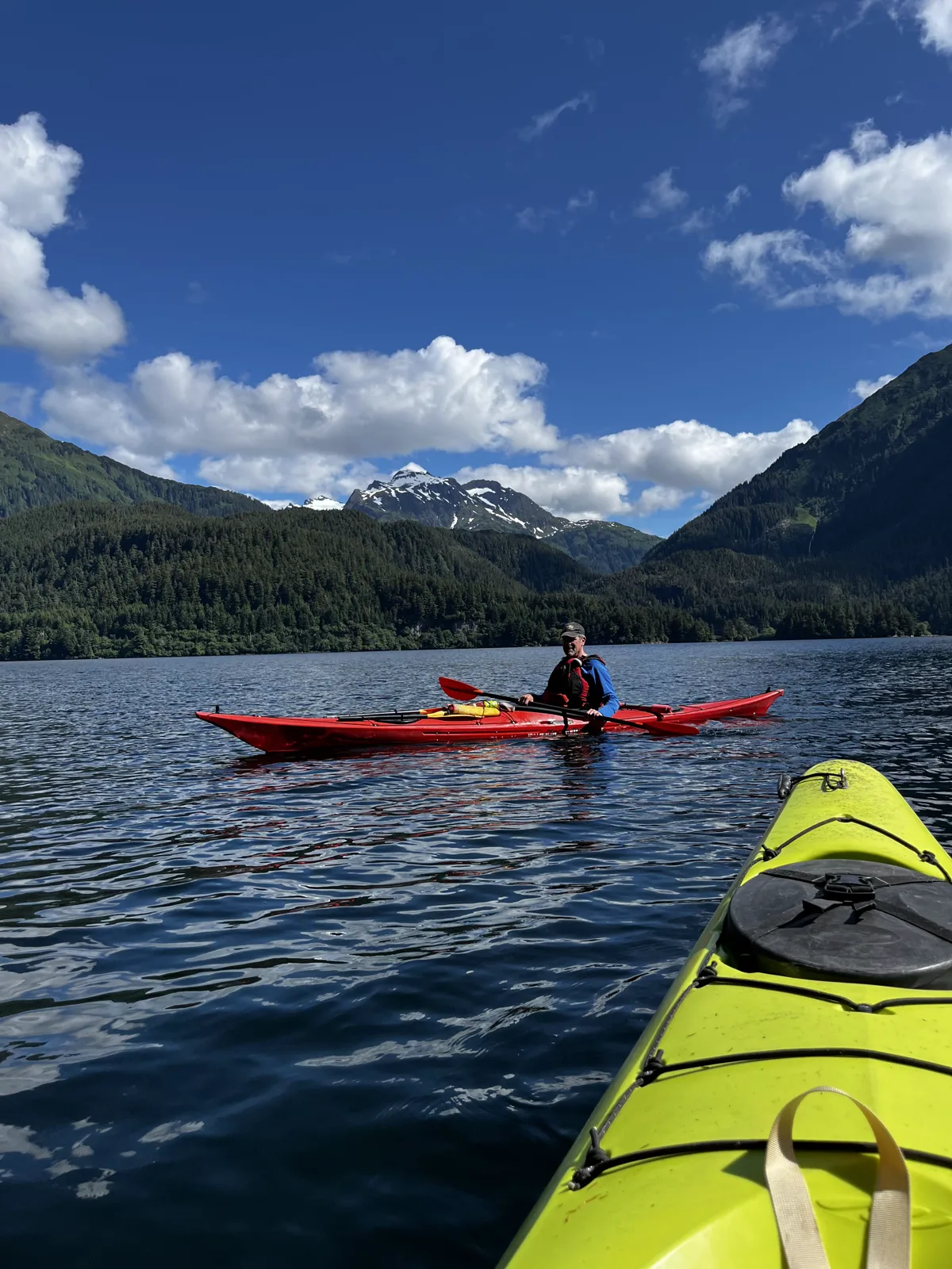 A paddler in a red sea kayak on calm water below snow-capped mountains near Sitka, Alaska